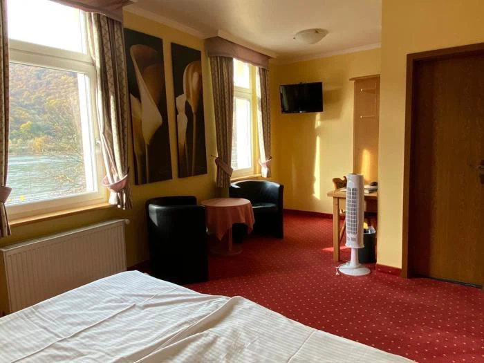 Et hyggeligt hotelværelse med to stole og et rundt bord ved store vinduer, en seng i forgrunden, tv monteret på væggen og rødt gulvtæppe.